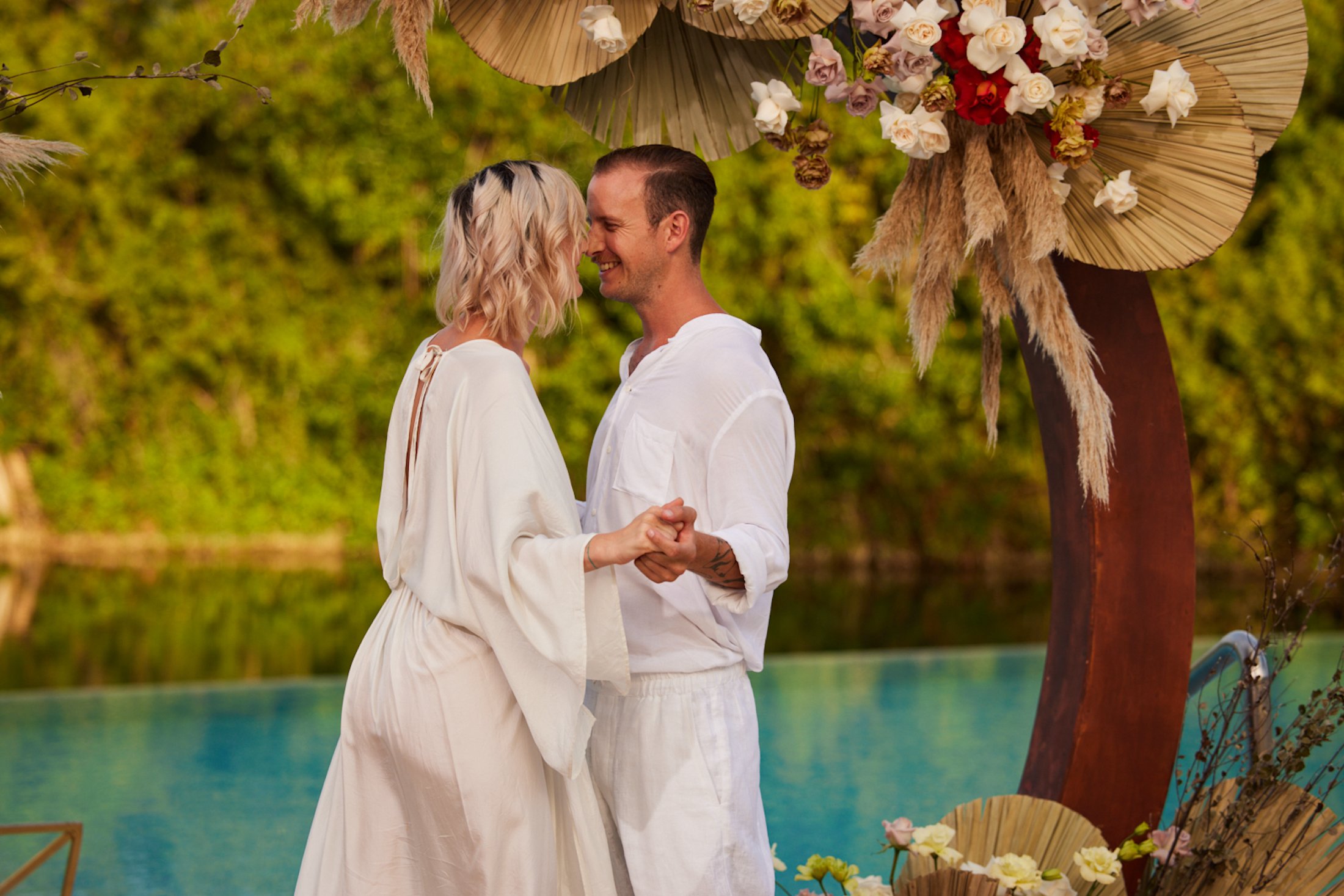 A man and woman in white canoodling under a flowery archway next to a river.