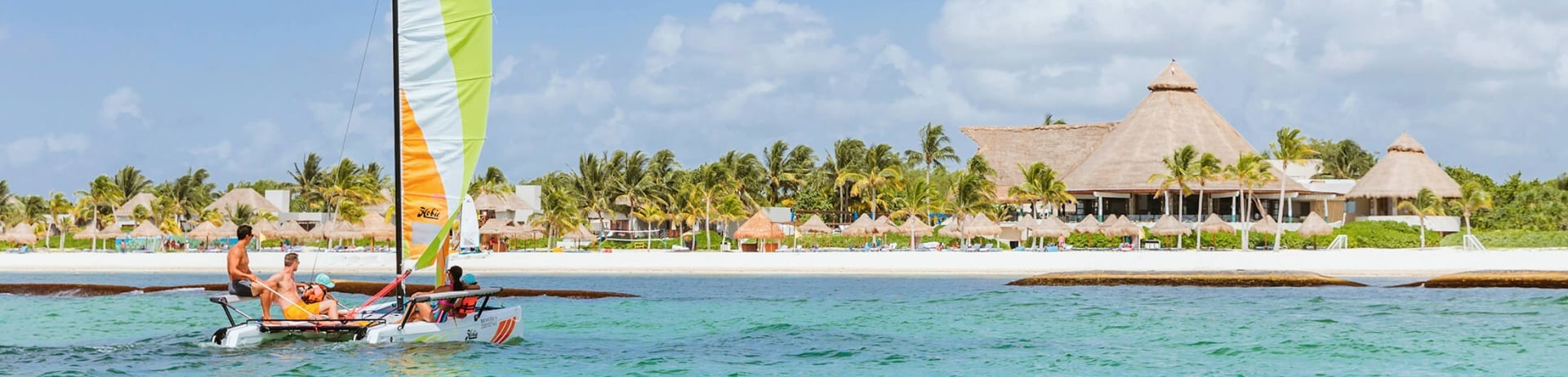 A group of people on a mini catamaran heading toward a white sand beach and a resort.