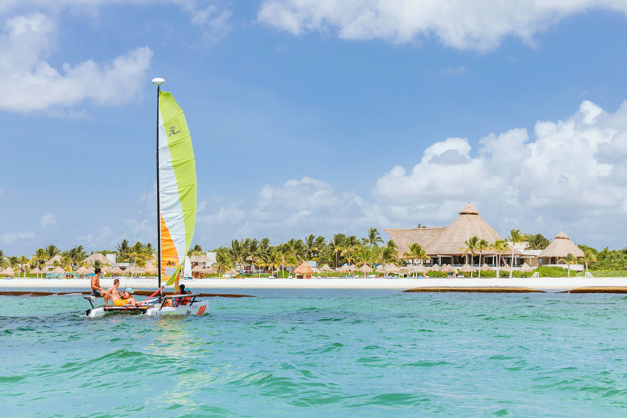 A group of people on a mini catamaran heading toward a white sand beach and a resort.