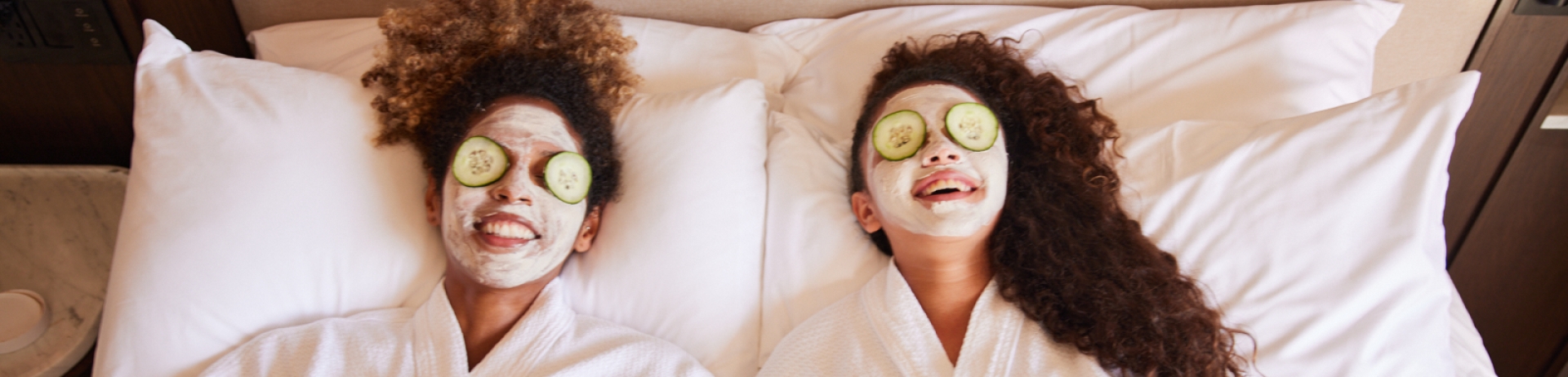 Two women lying in bed with robes and cucumbers on eyes