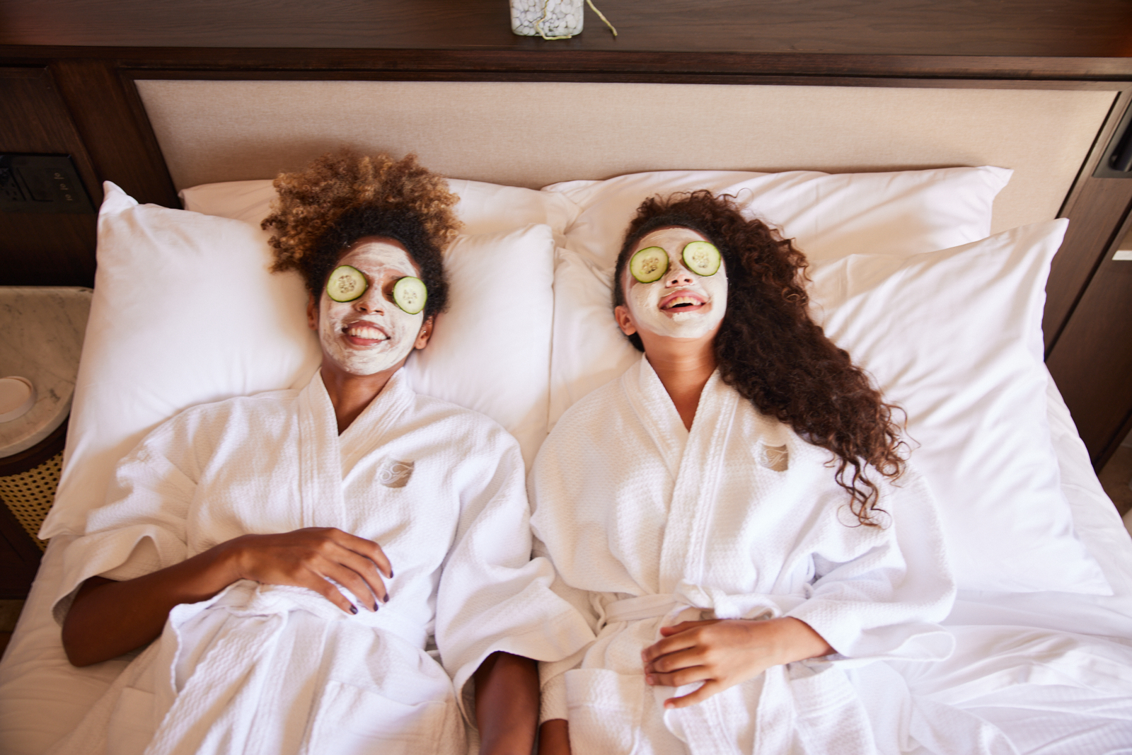 Two women lying in bed with robes and cucumbers on eyes